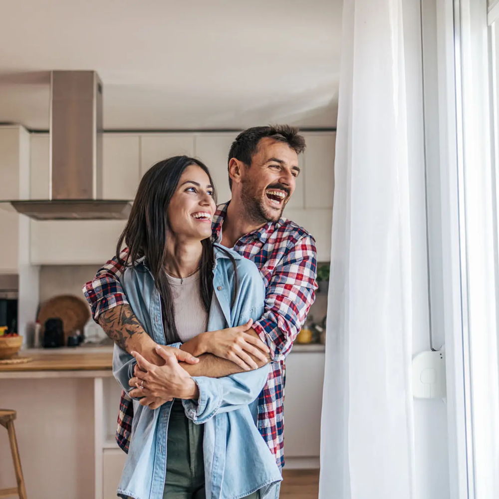 A happy couple embraces while looking out a window in a bright room.
